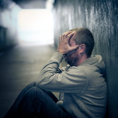 Profile view of a young homeless caucasian male - in his late 20s or early 30s - sitting in a dark, damp subway tunnel, his knees drawn up and his hands covering his face in desperation and despair. He is hungry, lonely and desperate. The man has short cropped hair and an unkempt beard. There is a look of sadness, loneliness and desperation in his eyes. He is sitting on a piece of old cardboard Horizontal image with room for copy space.